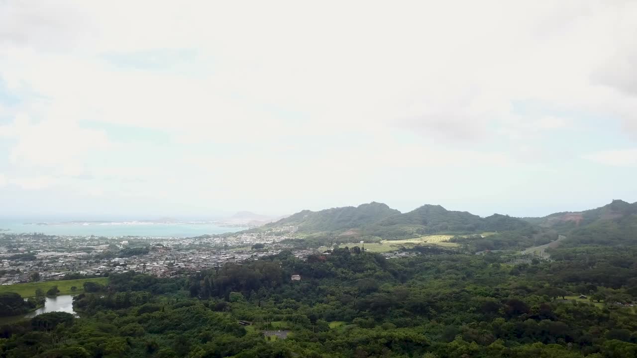 Wide aerial slow push-in from above Ho‘omaluhia area on Oʻahu’s Windward side, toward Kaneohe Valley with suburban homes, lush green hills, and Kaneohe Bay in the distance under bright hazy daylight