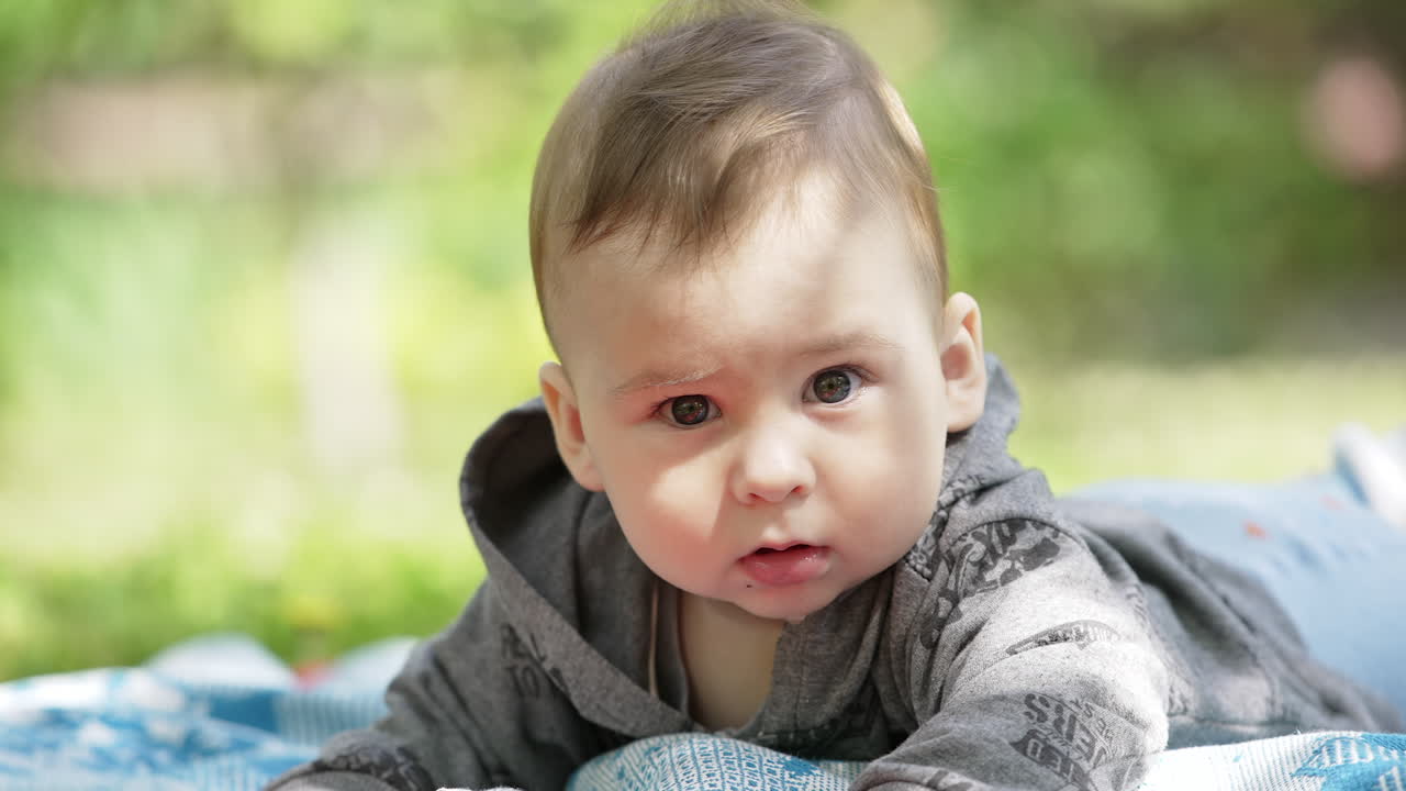 Healthy Caucasian kid in grey hoody lies in nature. Beautiful kid hits the ground with his arms. Close up.