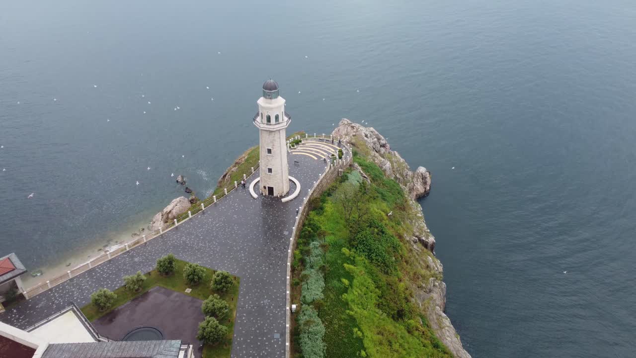 White lighthouse stands on narrow rocky peninsula surrounded by calm blue sea, lush greenery and paved path, elevated aerial perspective at Start Point Lighthouse, Devon, England