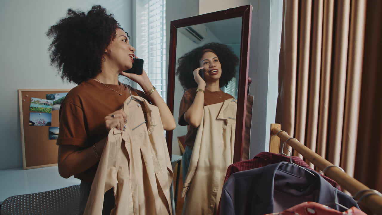 Curly Woman Talking on Phone while Trying on Beige Coat near Mirror