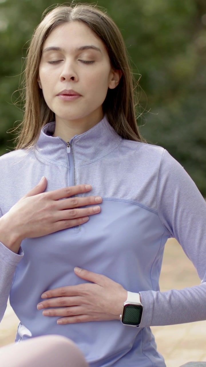 Video verticale: Donna che posa le mani sul busto e respira profondamente al parco, con top lilla e smartwatch per calmarsi.
