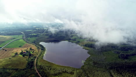 Overview-of-a-small-lake-by-the-countryside-surrounded-by-roads,-agricultural-fields-and-grasslands-all-around-on-a-cloudy-day