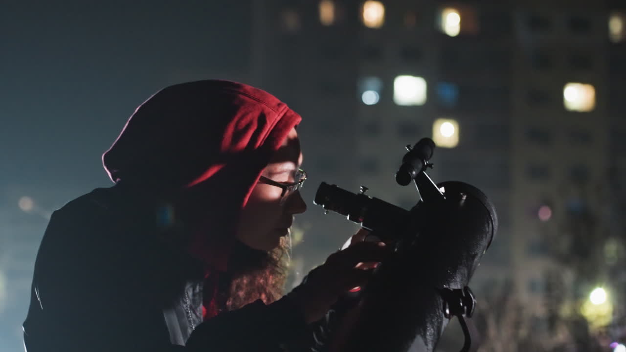 Un astronomo con cappuccio scruta attraverso il telescopio lo skyline notturno della città; un osservatore barbuto con una felpa rossa regola il telescopio sul tetto; bokeh delicato dalle finestre degli appartamenti; atmosfera tranquilla di curiosità e scientificità