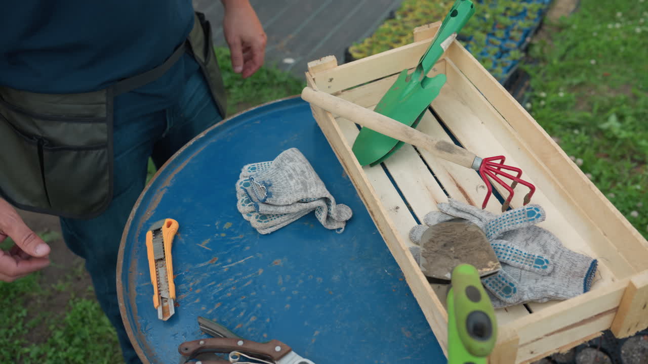 Male gardener wearing pocket apron picks hand shovel from wooden crate on blue metal drum and slips tool into side pocket, with gloves, pruners, utility knife, outdoor nursery setup