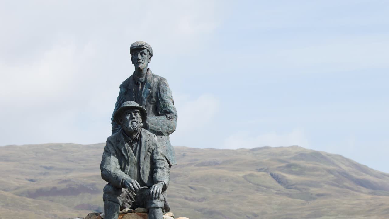 Bronze sculpture of two figures, outdoor daylight, mountains, brief person passing, static camera