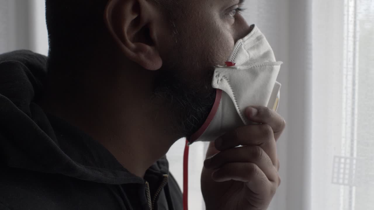Adult Asian Male Holding Up Wearing FFP Three Mask To Face Indoors Looking Through Curtain