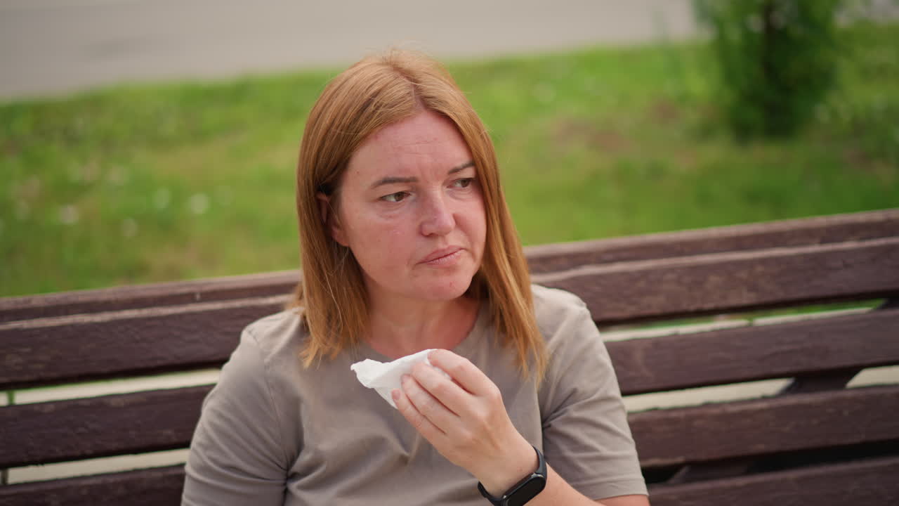 Crying woman sitting on wooden bench outdoors wiping tears with tissue under soft daylight, emotional facial expression showing sadness and reflection, green park background