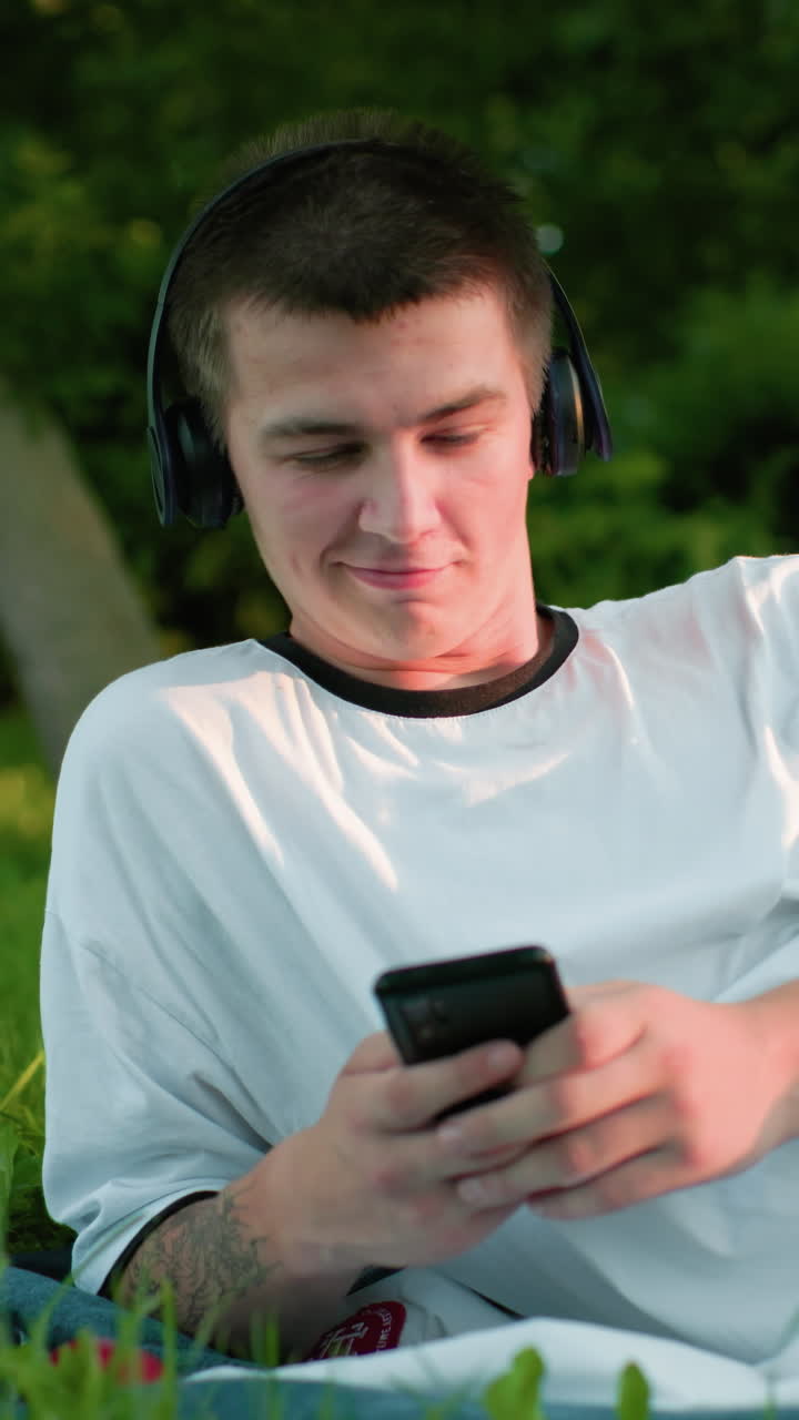 trabajador remoto con auriculares acostado en un campo cubierto de hierba con el teléfono en la mano, asintiendo a la música con una sonrisa sutil, rodeado de árboles y vegetación vibrante bajo la suave luz del sol