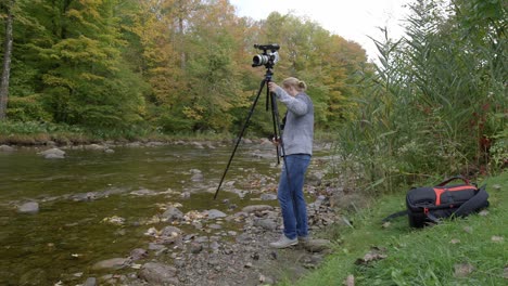 photographer adjusting tripod with video camera by flowing river, static shot