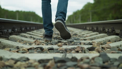 close-up view of a man's legs, dressed in jeans and canvas shoes, walking alone on rocky railway tracks with a blur view of trees and electric poles