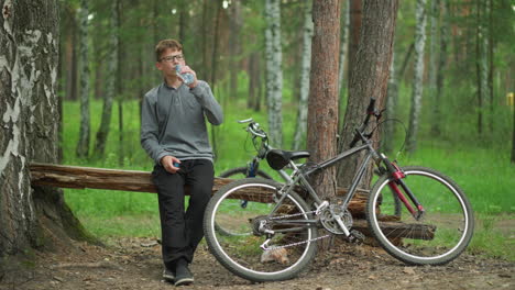 un niño joven con gafas y una camiseta de manga larga gris toma un sorbo de agua mientras está sentado en un tronco con la pierna cruzada, descansando al lado de una bicicleta estacionada en el bosque, rodeado de altos árboles verdes