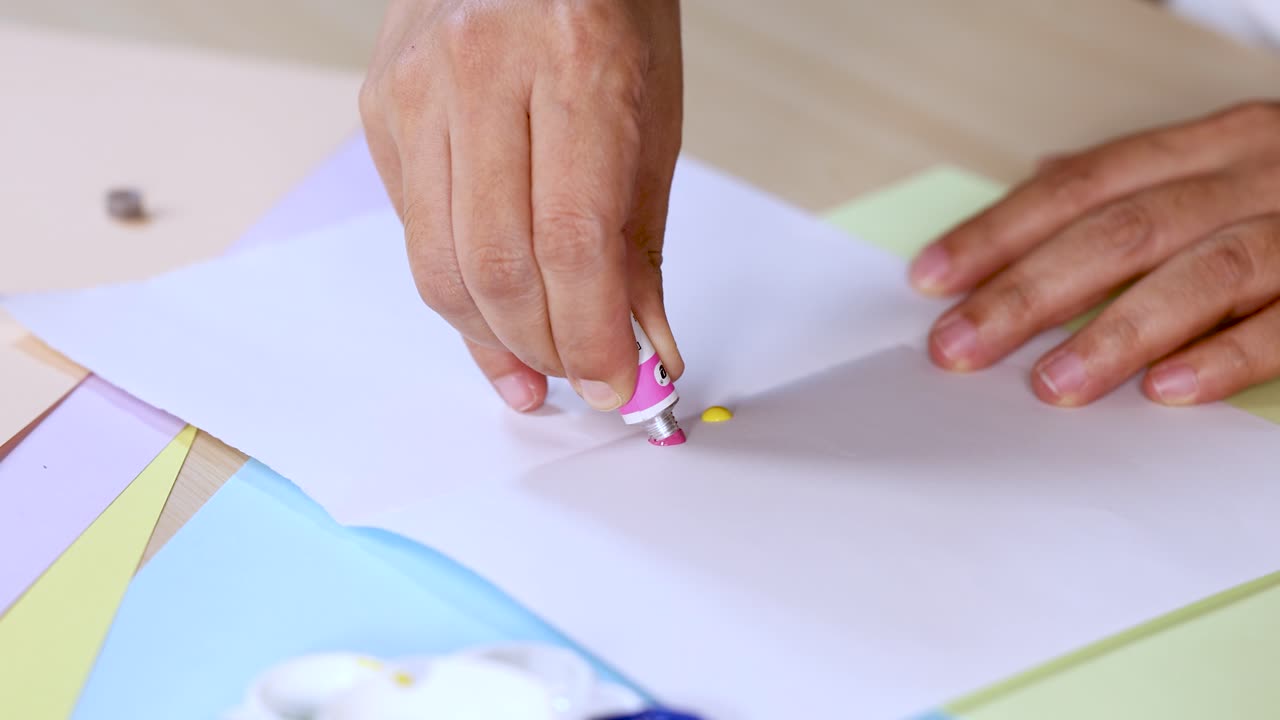 Hand squeezes pink and yellow paint onto white paper in bright, indoor art workspace