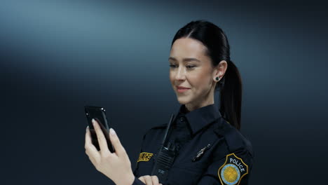 young joyful pretty policewoman in uniform smiling and having videochat on the smartphone via web cam on the dark wall background