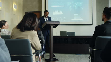 zoom de la caméra sur un homme d'affaires afro-américain parlant sur un podium dans une salle de conférence et montrant des tableaux et des graphiques sur grand écran