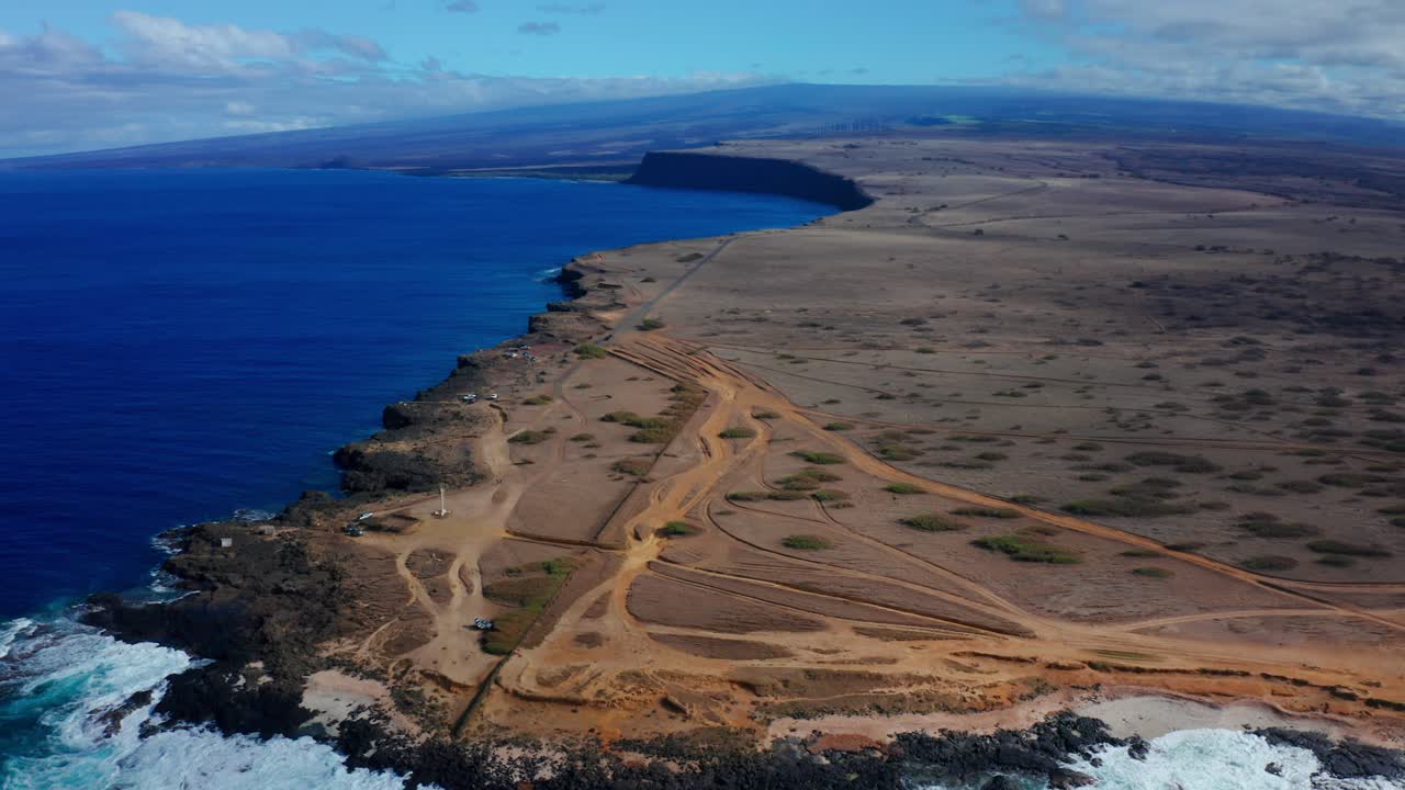 Endless rocky desert cliffs meet foaming blue waves as dusty tracks cut across the barren land, all under a bright sky and distant blue horizon.