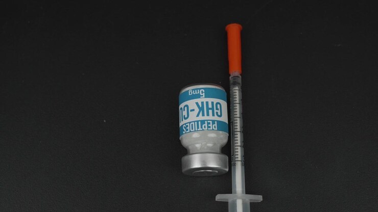 Medical insulin syringe rests across a labeled GHK-Cu peptide vial against a dark studio background, useful for healthcare, biotechnology, or pharmaceutical themes.