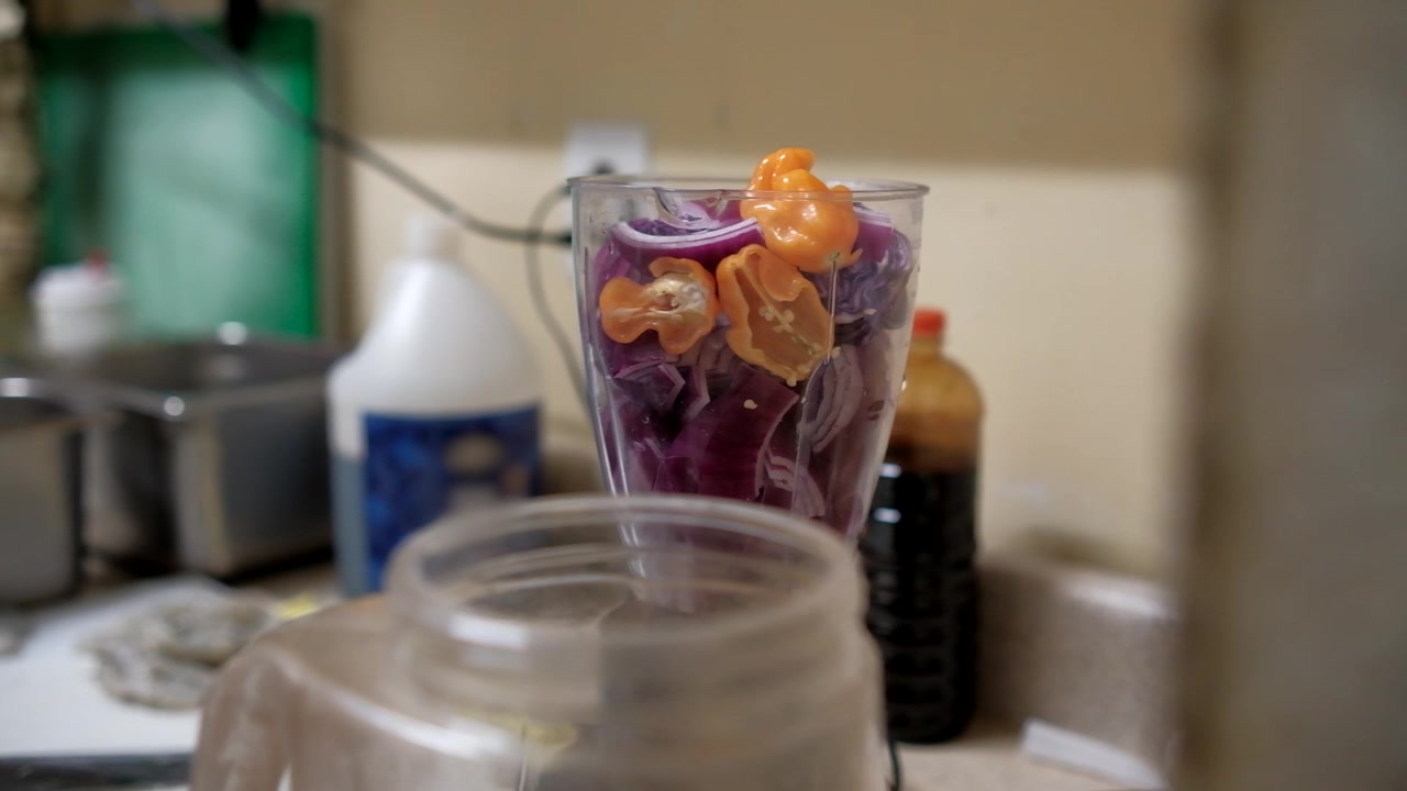 Vertikal närbild av orange habaneropeppar, lila rödlök och mörk vätska i lager inuti en genomskinlig mixerkopp innan man gör en kryddig sås för mexikansk räkaguachile