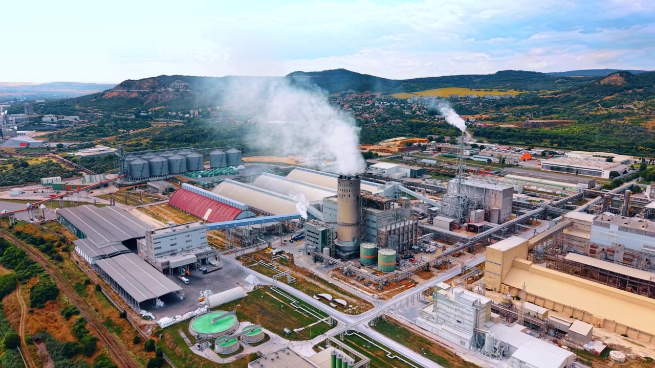 Büyük bir endüstriyel işleme tesisinin üzerinde yükselen buhar bulutu. Ağaçlık tepelerin altında fabrika holleri, tanklar, boru hatları ve demiryolları ön planı dolduruyor.