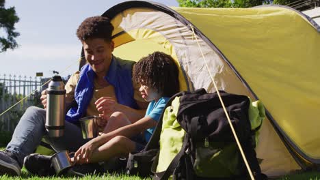 Happy-biracial-man-and-his-son-drinking-from-thermos-flask-in-garden