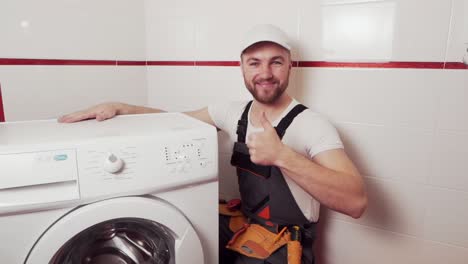 fontanero con uniforme de trabajo conectó la lavadora y muestra con el pulgar hacia arriba que todo está bien