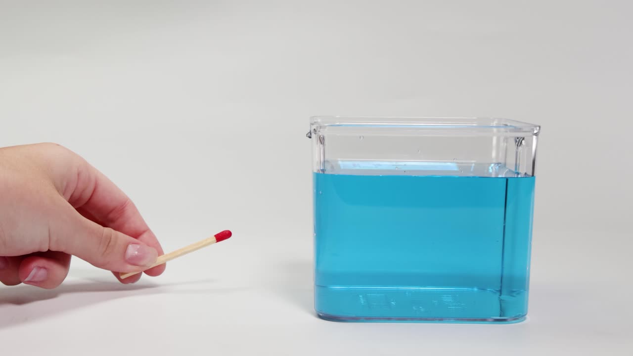 A hand places a matchstick in blue liquid, observing its buoyancy and movement in a clear container