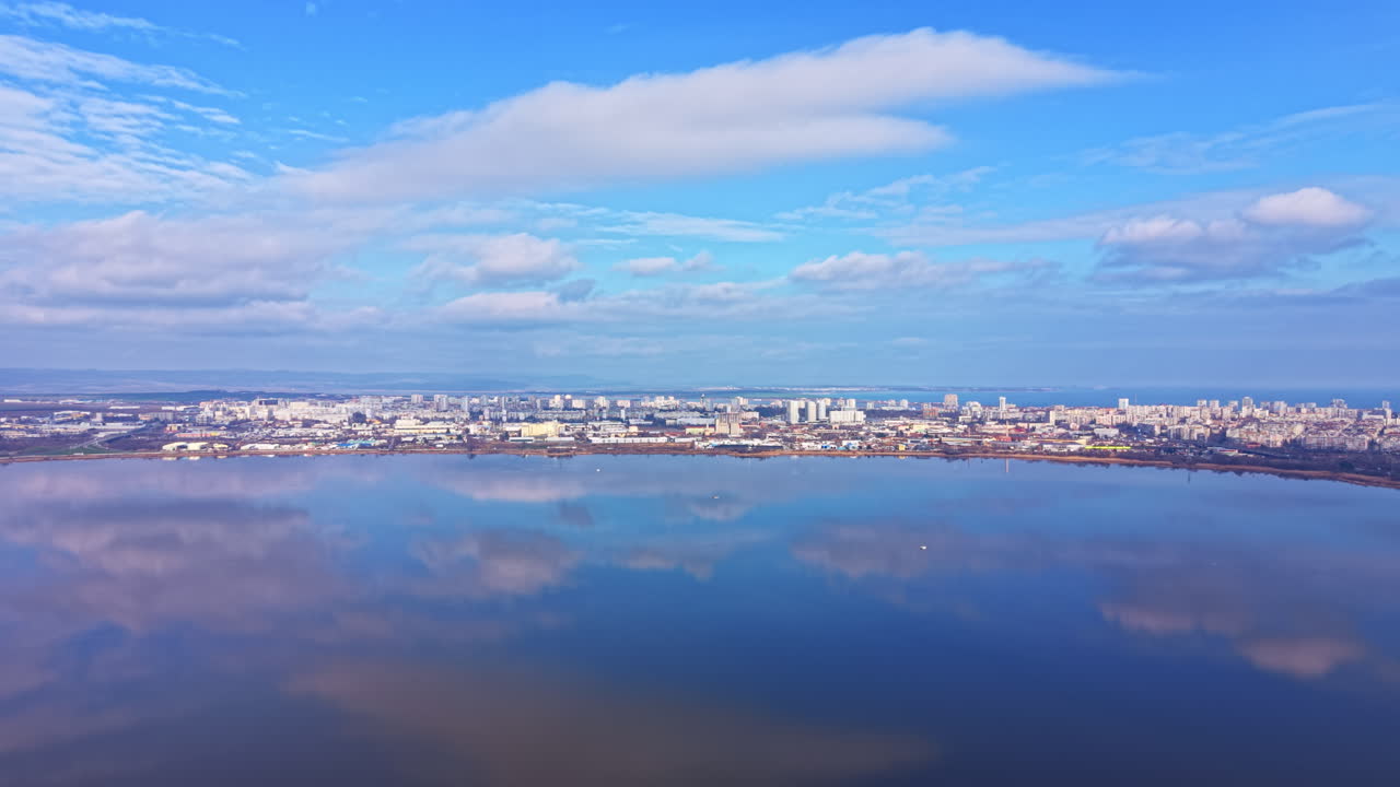 日中の水面に映る都市のスカイライン、空には雲、岸辺には建物が立ち並ぶ景色
