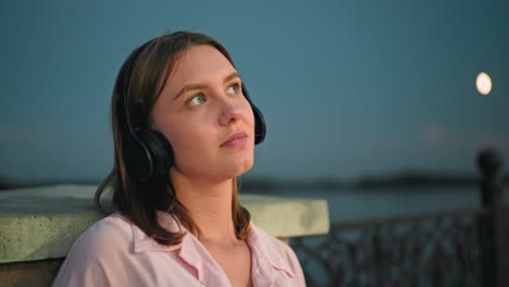 young lady in pink top and headphones resting on fence looking up thoughtfully with small insect flying close to her face, moon in background