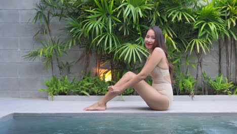 A-young-attractive-Asian-woman-with-long-brown-hair-and-in-a-one-piece-bathing-suit-sits-along-the-side-of-a-pool