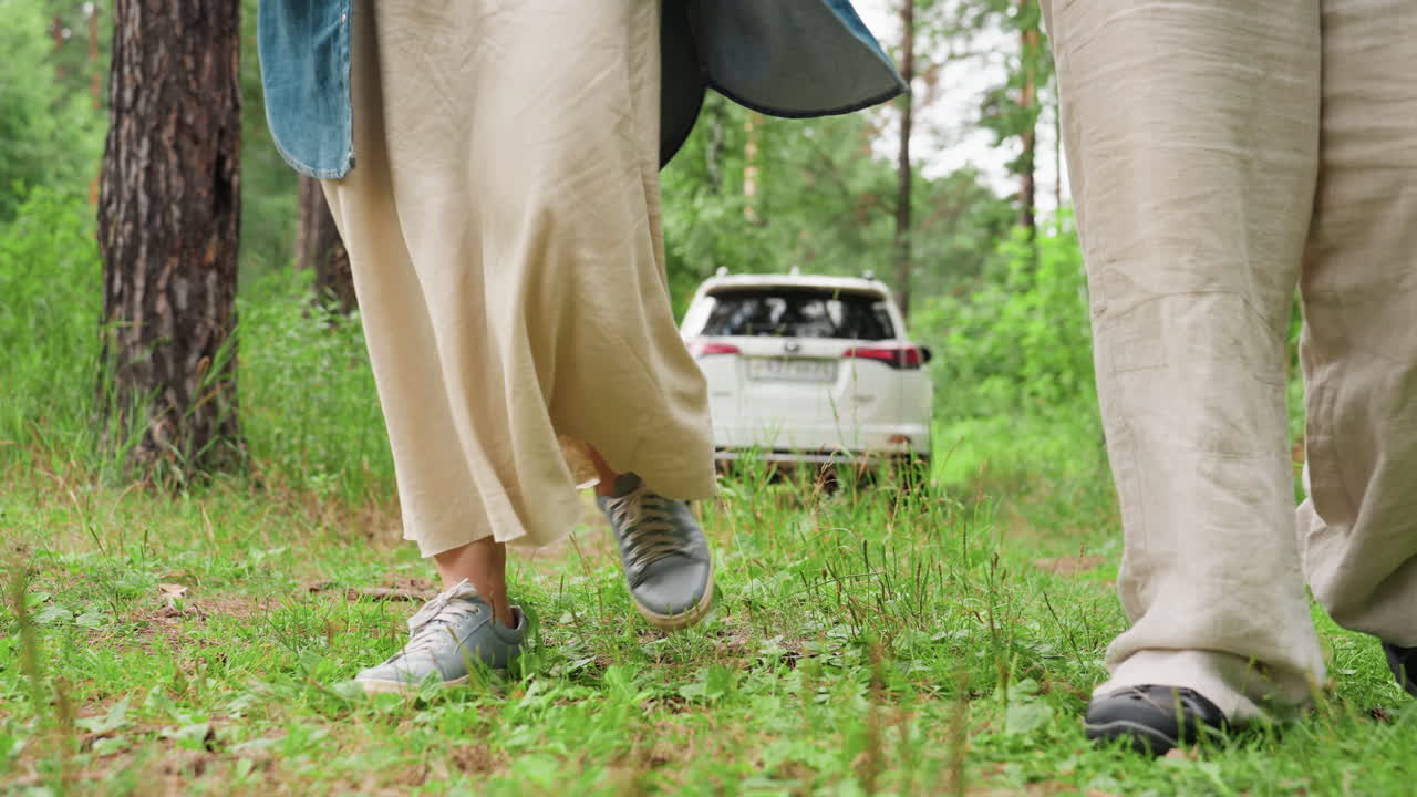 사랑스러운 커플이 손을 잡고 주차된 차에서 평화로운 숲길을 걸어가며 맞춰 입은 옷을 입고, 울창한 녹색과 자연광으로 둘러싸여 함께 차분한 산책을 즐기고 있어요.