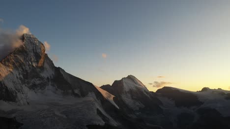 Toma-Aérea-Panorámica-De-Derecha-A-Izquierda-De-La-Montaña-Matterhorn,-Suiza,-Durante-Un-Amanecer-Claro,-Tranquilo-Y-Silencioso-Con-Un-Cielo-Azul-Y-Una-Pequeña-Nube-Delgada-Que-Rodea-El-Pico-Nevado-Durante-El-Verano
