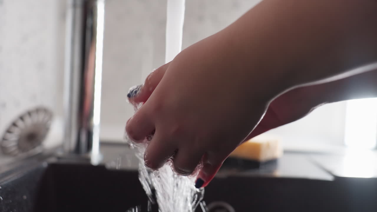 Nærbillede af hænder, der vasker et æble grundigt under rindende vand i køkkenvasken. Blå manicure er synlig, skum og vandstænk, omhyggelig skylning for hygiejne, klargøring til ren spisning med vandhanestrøm.