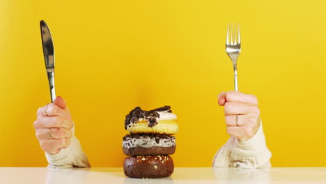 two hands holding a knife and fork eagerly in front of a stack of three donuts against a bold yellow background, creating a playful and appetizing dessert scene.