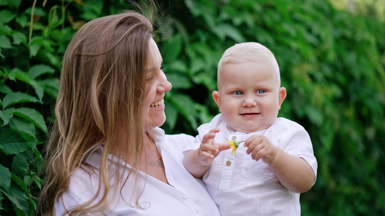 Attractive long-haired Caucasian woman holding a cute blond baby boy. Happy mother with her beloved son holding a flower in hand.
