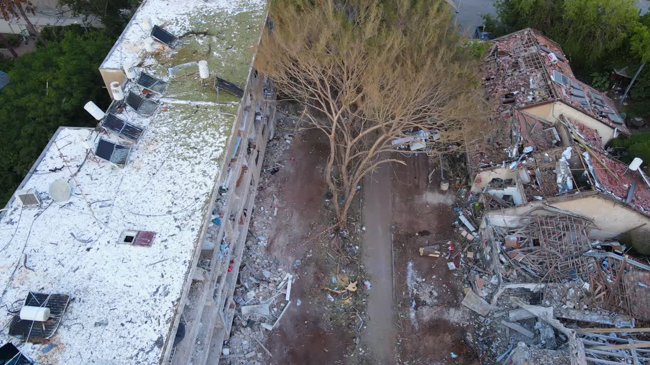 Top-down drone shot of a devastated neighborhood in Tel Aviv. A tree lies in the center, surrounded by ruins and destroyed buildings on both sides