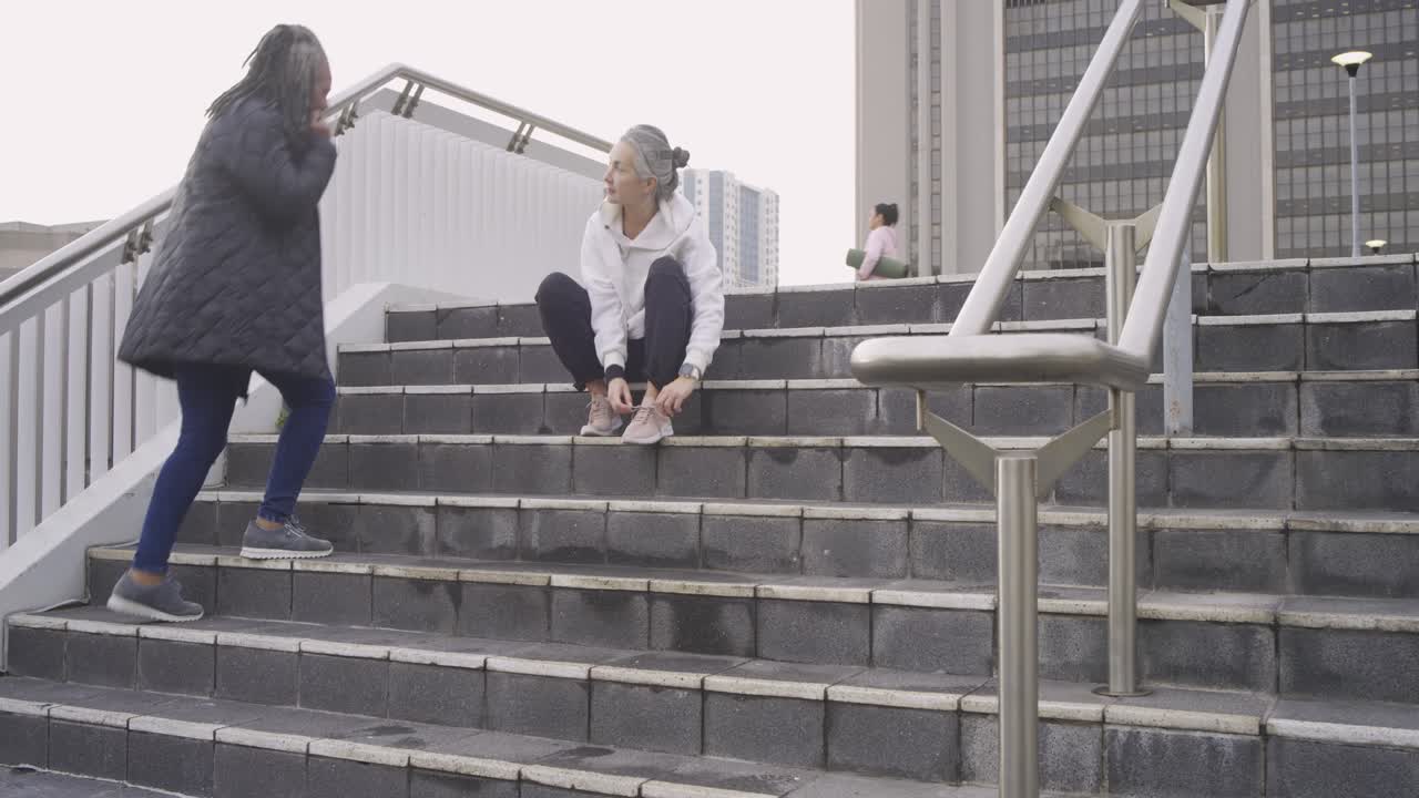 Mujer blanca madura abrochándose los cordones rosas de sus zapatillas en unas escaleras de piedra antes de correr, revisando su reloj deportivo.