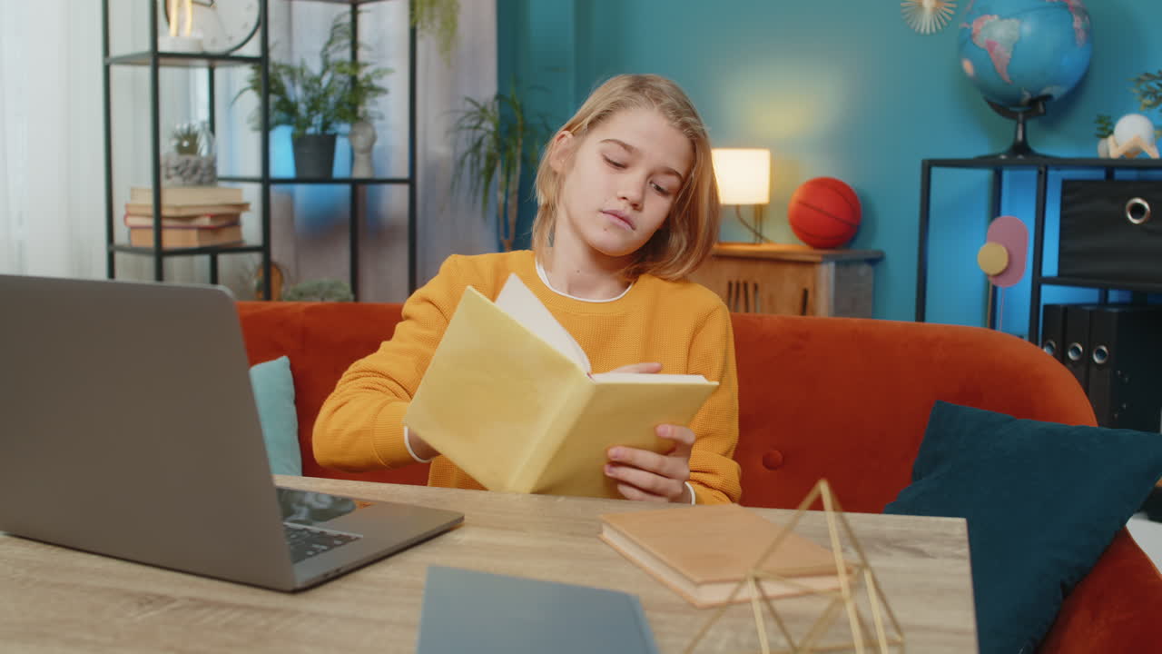 Teenager boy doing online homework on laptop reading book and taking notes on couch at home room