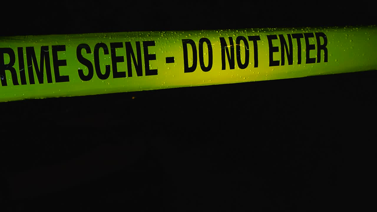 Close up view of a police forensics investigation yellow colored crime scene tape pulled in front of a pitch black night area while raindrops are falling on the tape. Ribbon is illuminated well