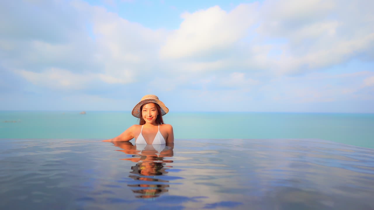 얼굴에 햇빛이 비치는 카메라를 바라보는 고급 호텔 리조트의 인피니티 풀에 있는 아시아 여성, 풀 프레임