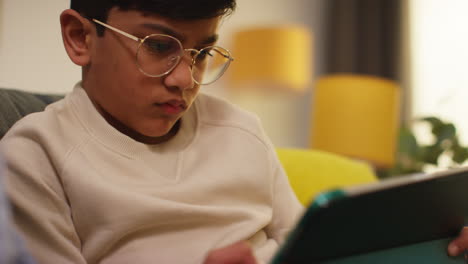 close up of young boy sitting on sofa at home playing games or streaming onto digital tablet 6