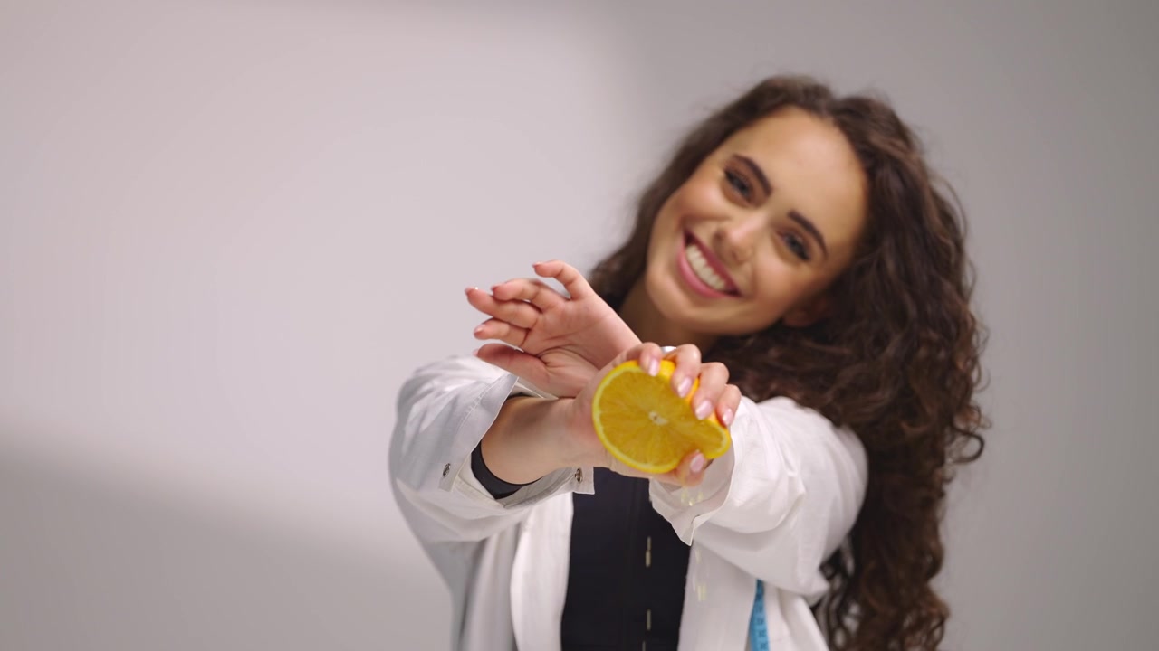 Giovane nutrizionista sorridente in camice bianco sprema mezza arancia fresca per il succo mentre guarda in camera, presentando frutta salutare su uno sfondo bianco pulito per benessere e nutrizione