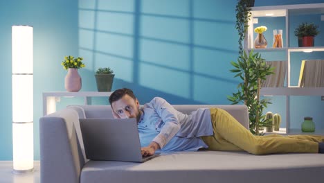 Happy and sympathetic man using laptop while lying on sofa at home.