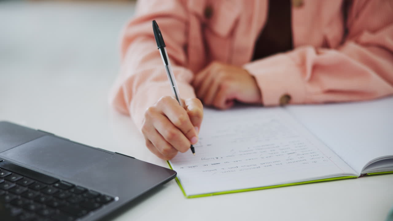 Person writing in a notebook with a pen next to a laptop