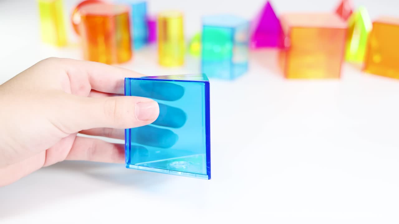 Hands manipulate colorful 3D geometric shapes on a white surface, highlighting prisms and pyramids in a bright setting