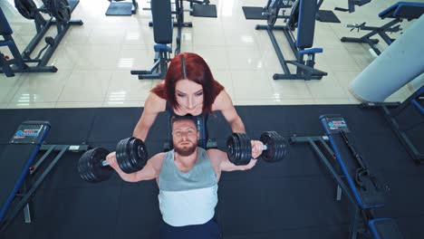 muscular bearded man is lifting heavy dumbbells on the gym background. attractive female instructor helps a strong athlete to lift dumbbells. top view.