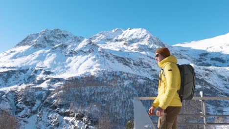 Young-caucasian-man-with-a-yellow-jacket,-beanie,-sunglasses-and-bagpack-walks-to-a-railing-and-takes-a-look-around-to-the-mountains-on-a-winter-sunny-day-in-Alp-Grum,-Switzerland