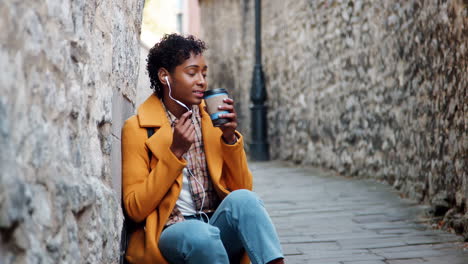 young woman wearing a yellow pea coat and blue jeans sitting in an alleyway in a historical city talking on her smartphone using earphones, close up