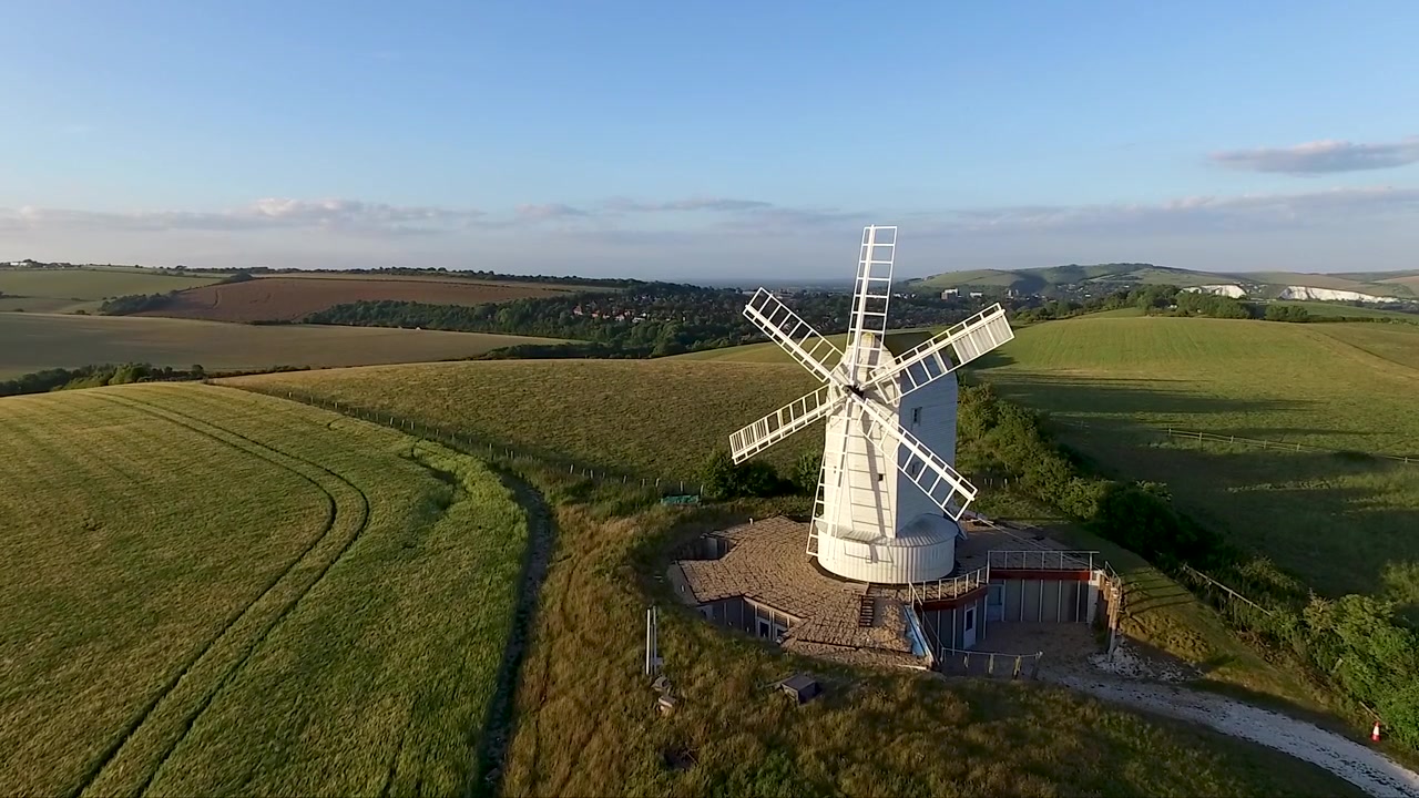 Aerial drone approach to a historic windmill, rising higher as it moves closer, revealing expansive green fields, rolling countryside and a scenic rural landscape in warm natural light