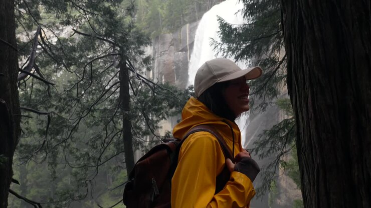 Happy Woman in Yellow Raincoat Looking at Majestic Waterfall in Forest