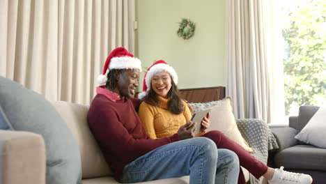 Feliz-Pareja-Diversa-Con-Sombreros-Navideños-Usando-Tableta-En-El-Sofá-En-Casa,-Espacio-Para-Copiar,-Cámara-Lenta