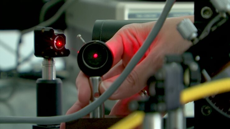 Laseropsætning i fotonisk laboratorium. Laserstråle. Hånd placerer linse for at rette laseren. Laserstrålen skinner på linsen.